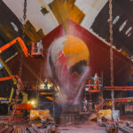 Workers use lift platforms and high-pressure equipment to clean and prepare the bow and bulbous bow of a large vessel inside a shipyard dry dock