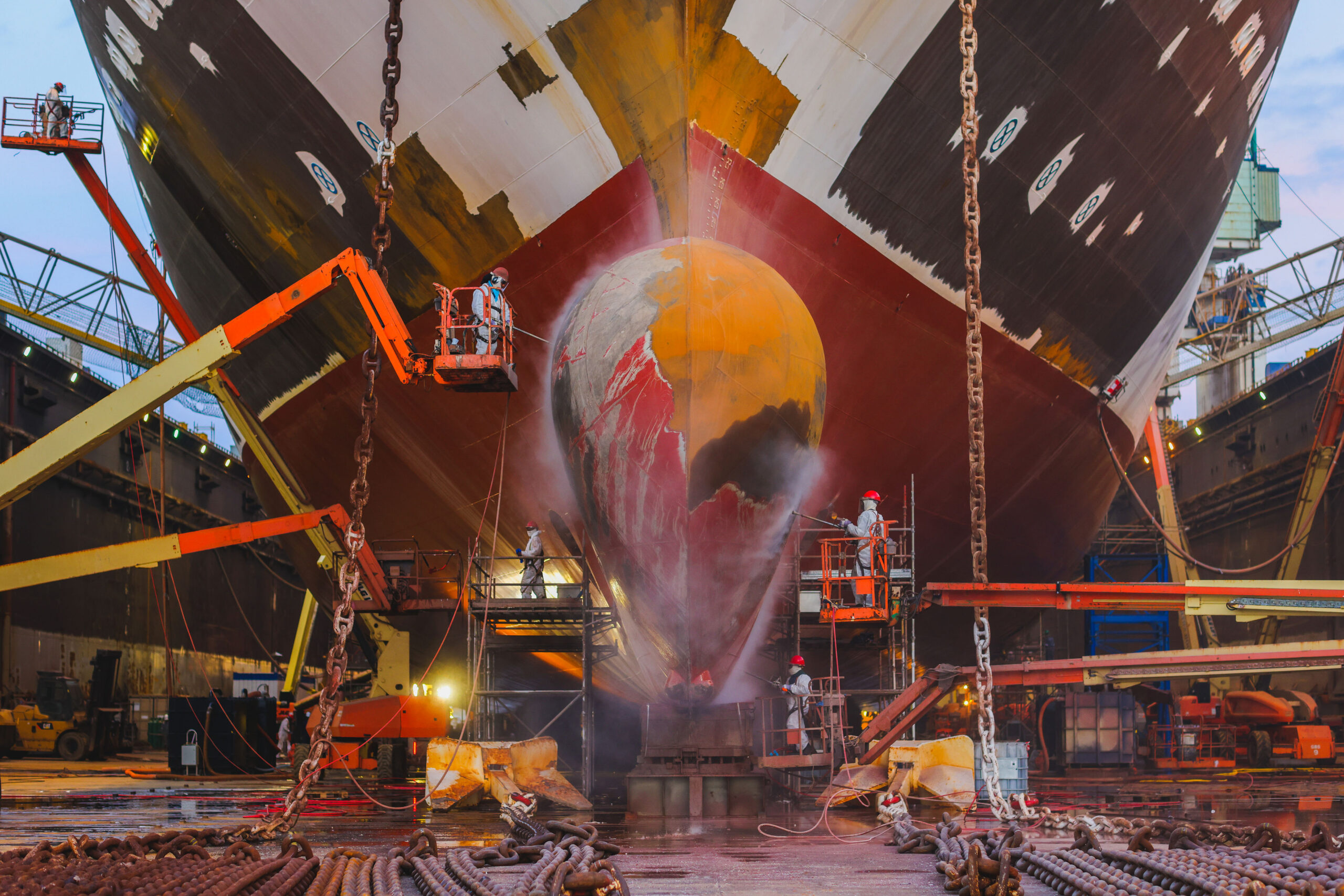 Workers use lift platforms and high-pressure equipment to clean and prepare the bow and bulbous bow of a large vessel inside a shipyard dry dock